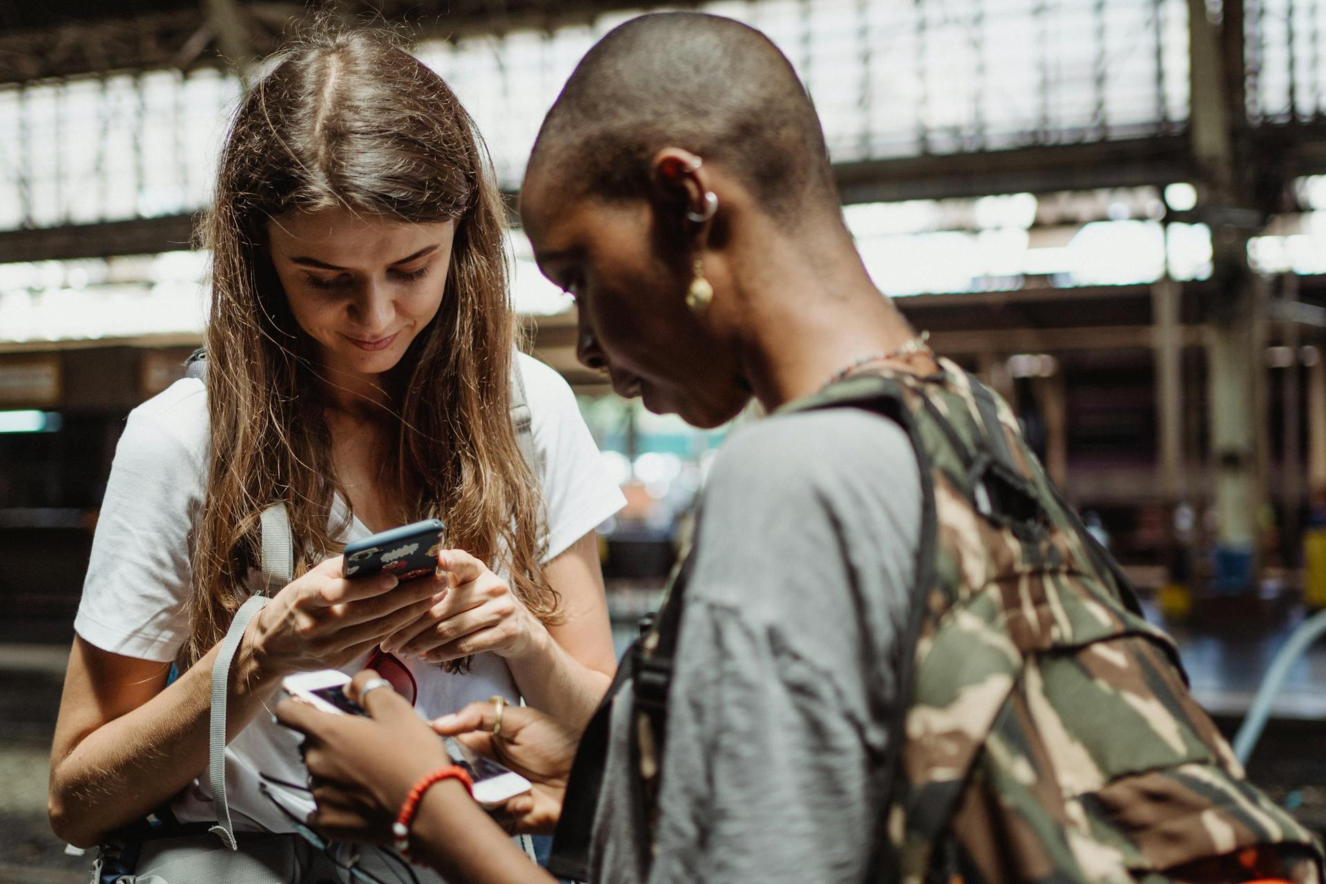 Two women using smartphones while standing in a busy train station, engaging in travel planning.