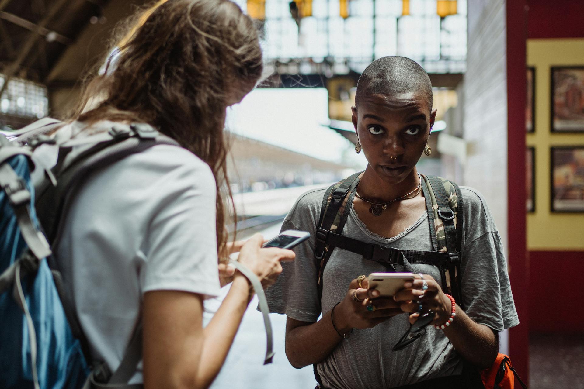 Two diverse female travelers using smartphones at a train station, backpacks visible.