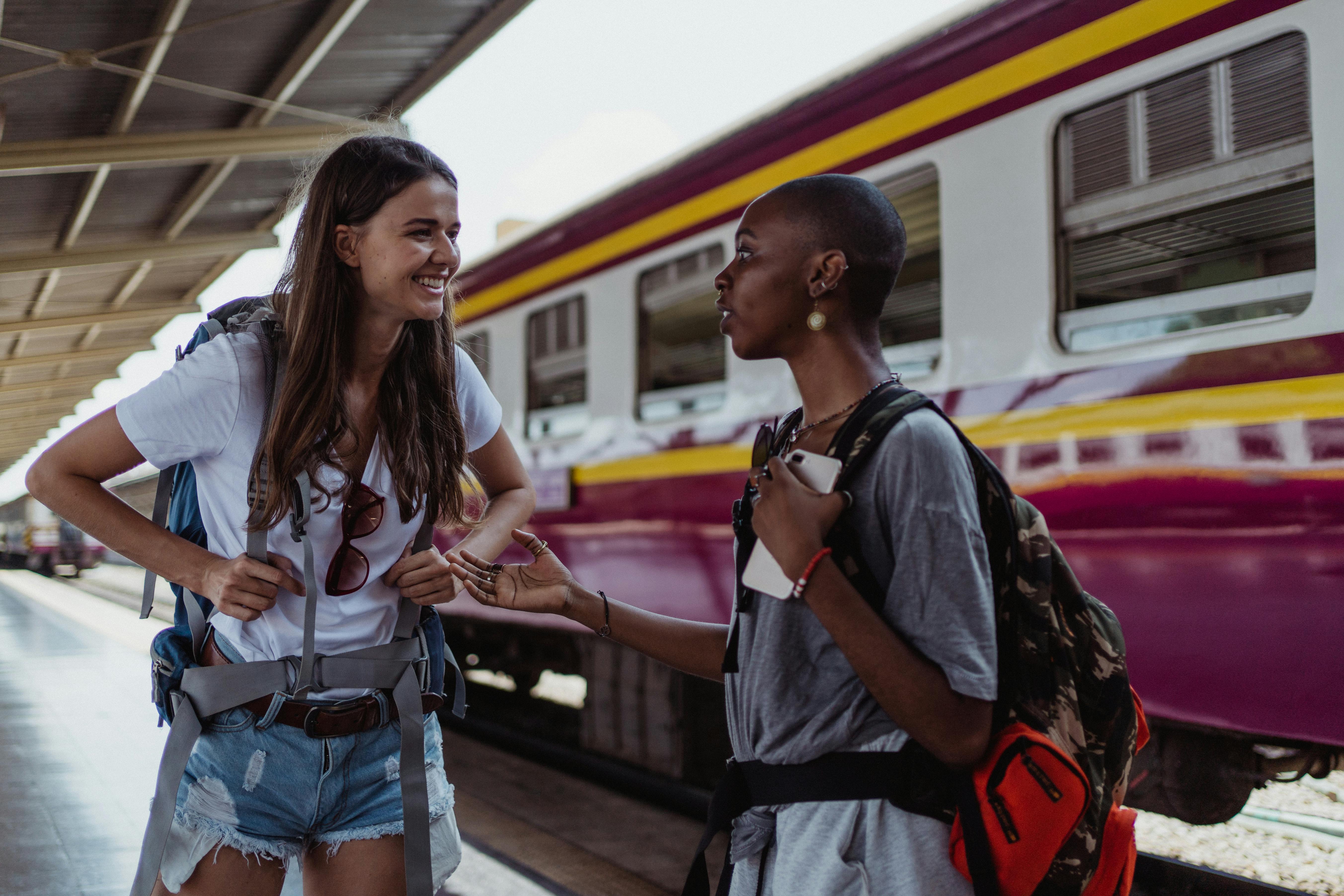 Two backpackers engage in friendly conversation at a railway station, with a train in the background.