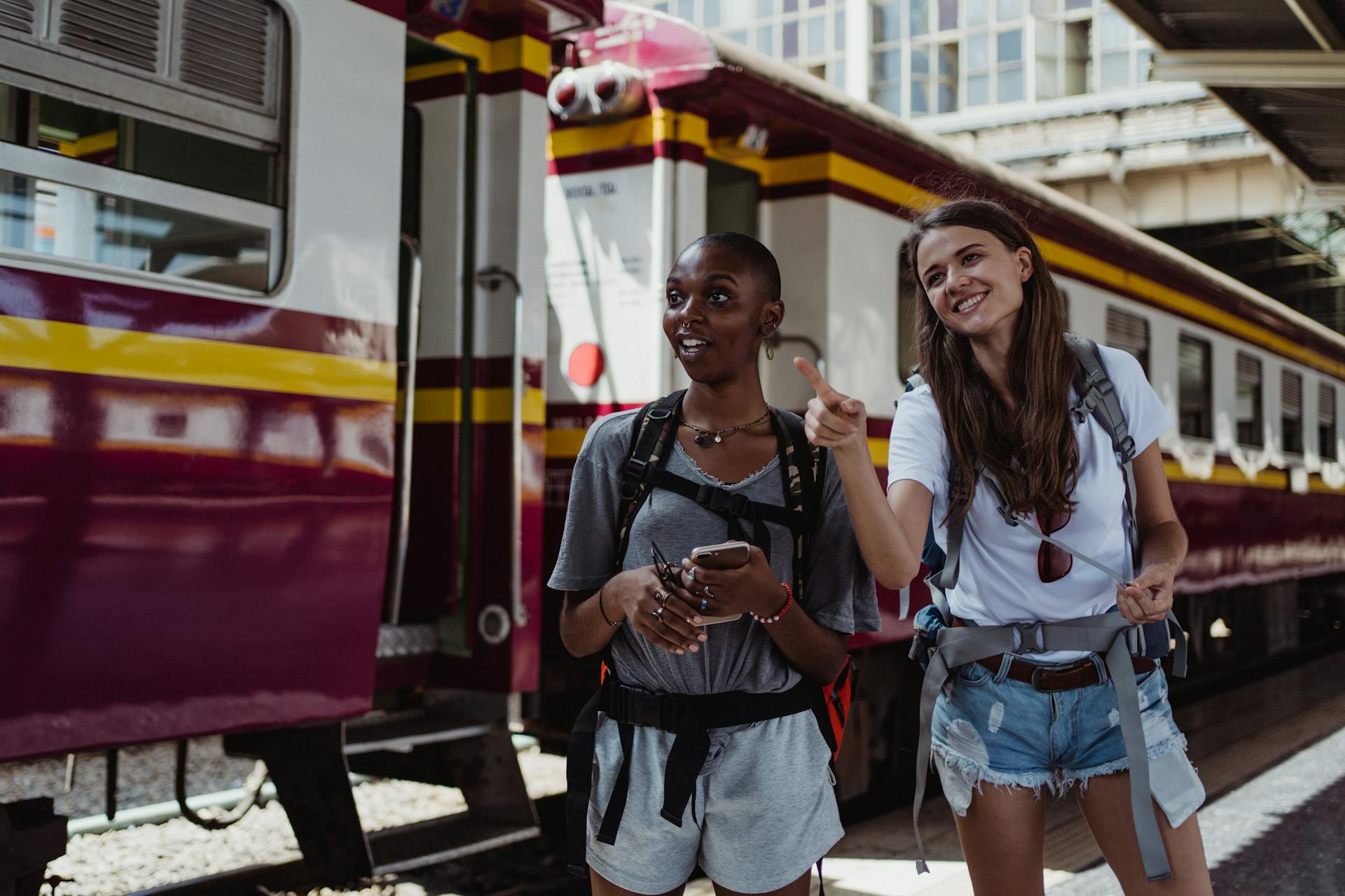 Two women with backpacks smiling and pointing at a city train station, ready for their travel adventure.