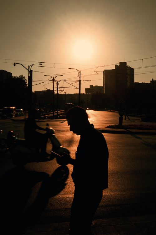 Free Silhouettes of a pedestrian and scooter rider against a city sunset backdrop. Vibrant urban street scene. Stock Photo