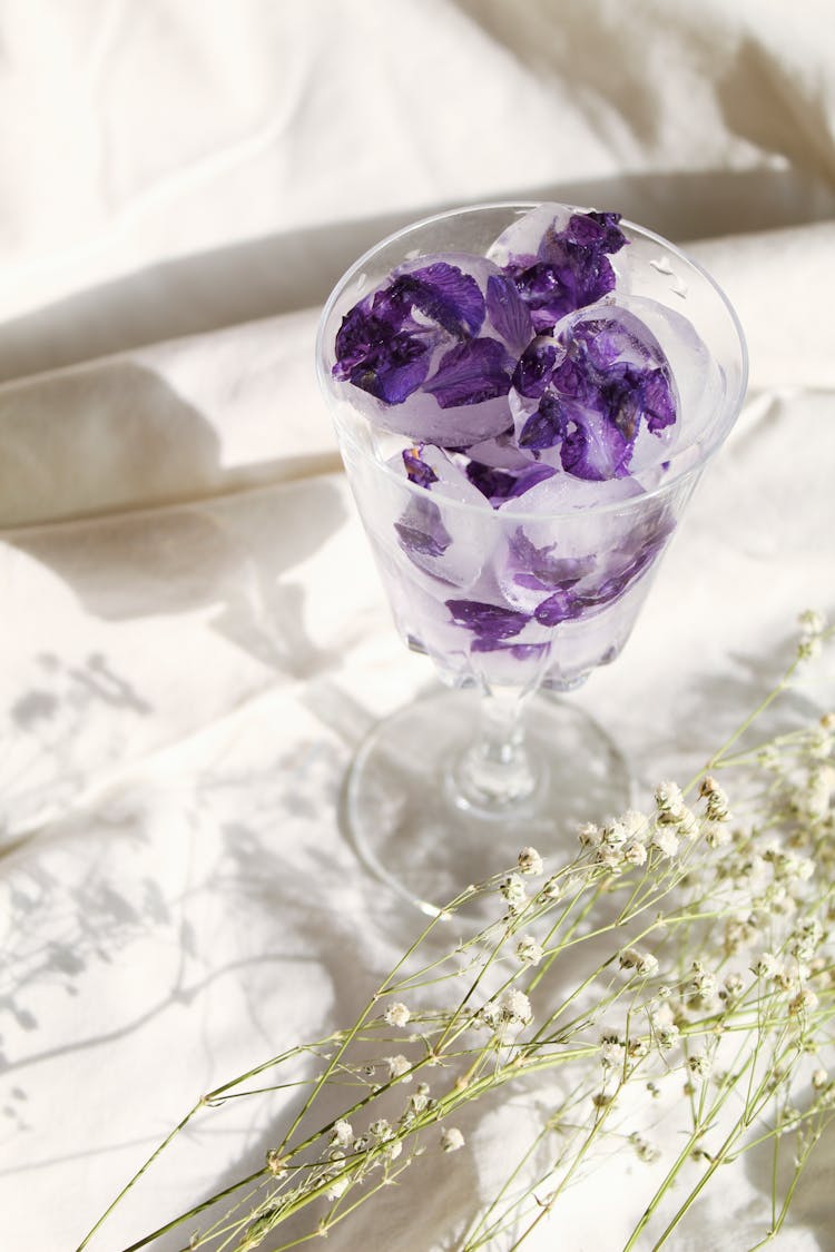 Purple Petals In A Glass With Ice And Flowers On Sheets In Sunlight