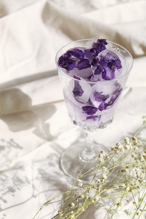 Purple Petals in a Glass with Ice and Flowers on Sheets in Sunlight