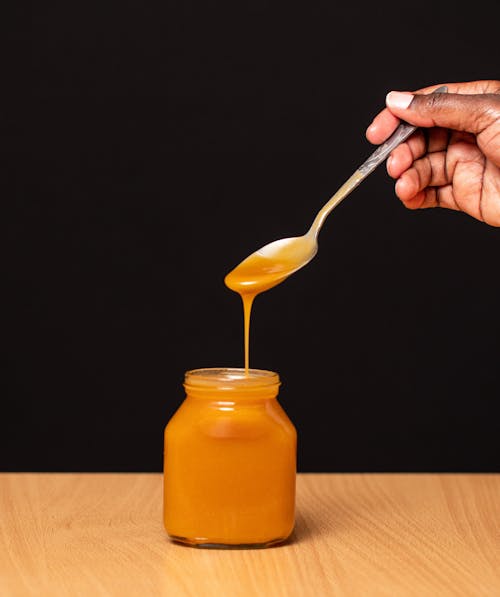 Person Holding Steel Spoon Above Honey Jar