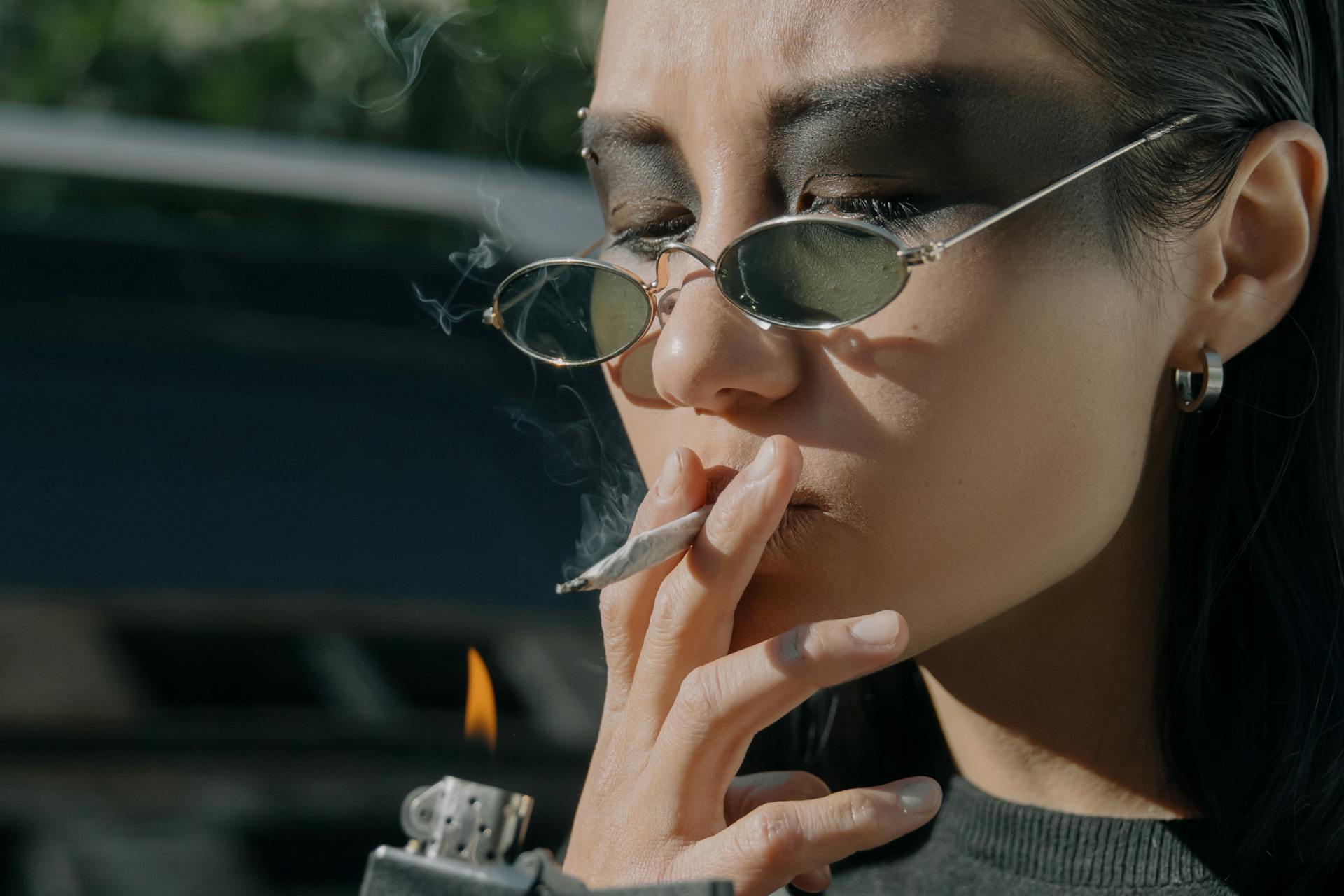 Portrait of a woman with dark makeup and eyewear smoking a cigarette outdoors.