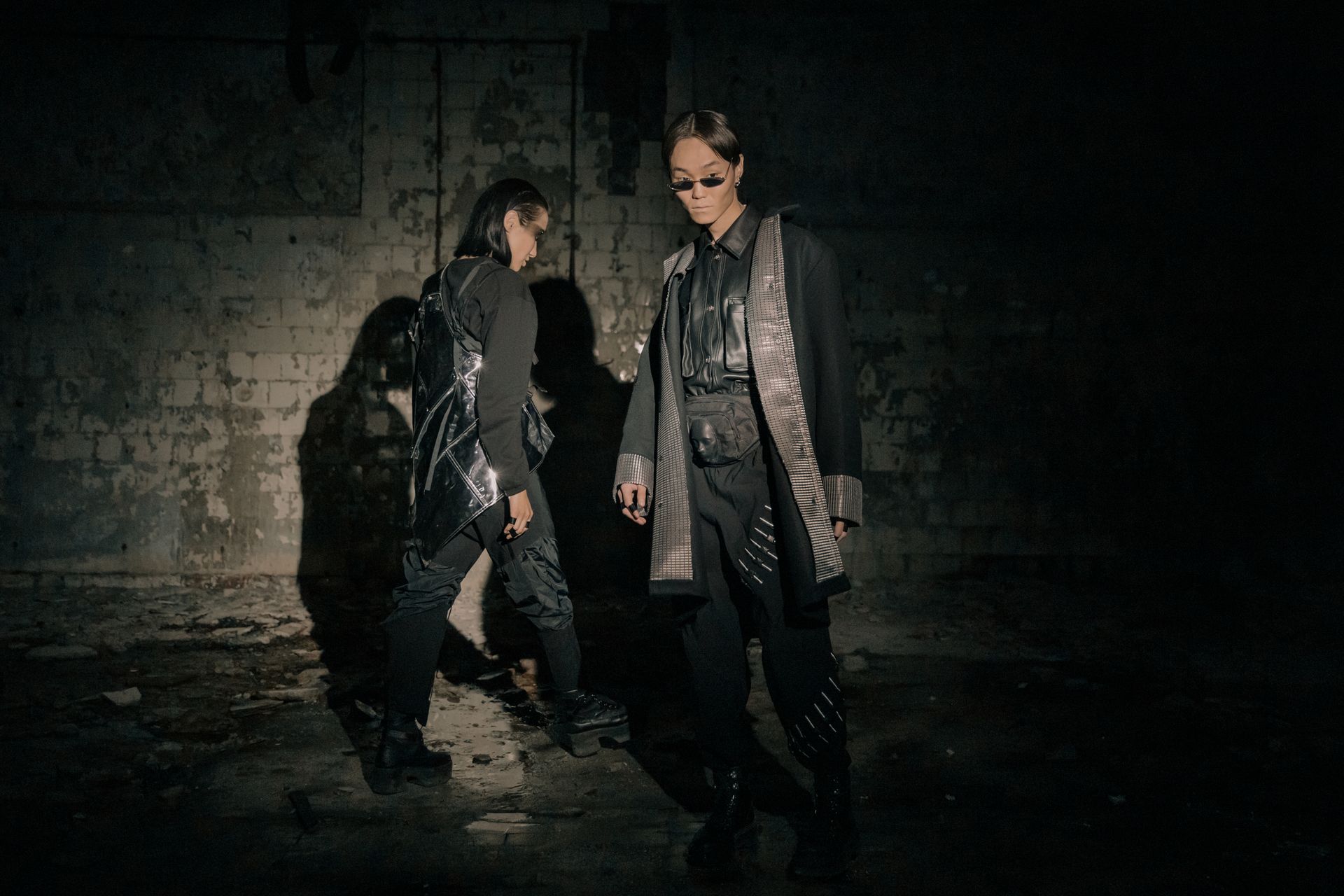 Two models in futuristic fashion pose in a dimly lit urban setting, exuding a cyberpunk vibe.