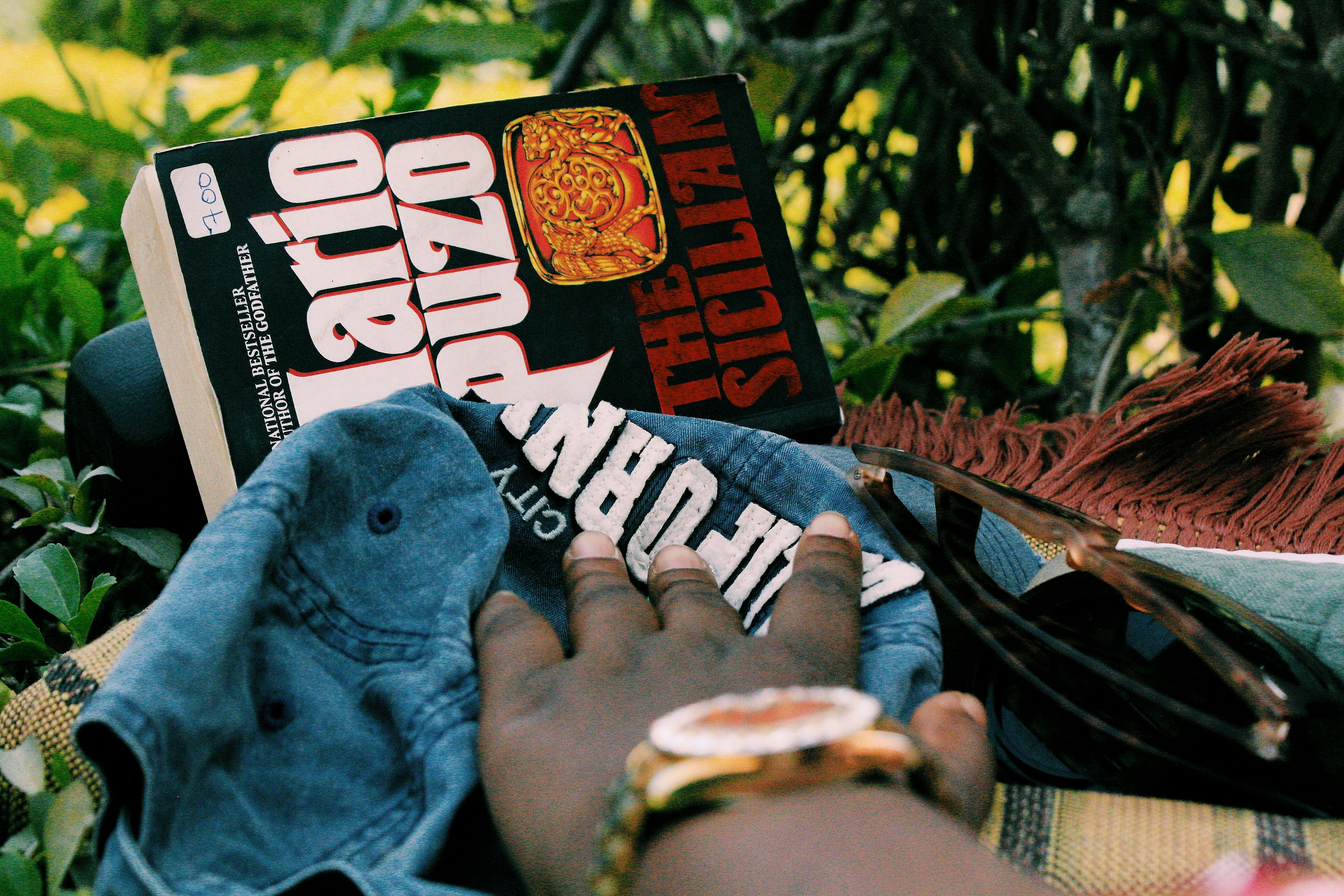 Faceless black person with golden watch sitting on grass with book near denim jacket and sunglasses in summertime in nature in daylight