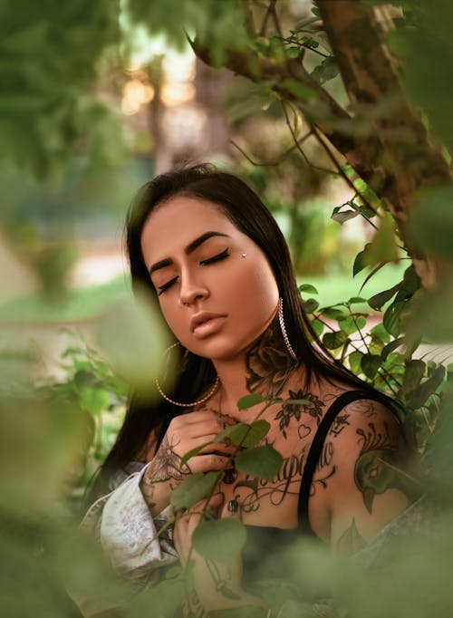 Young female with long dark hair and tattoos standing with closed eyes near green trees in daytime