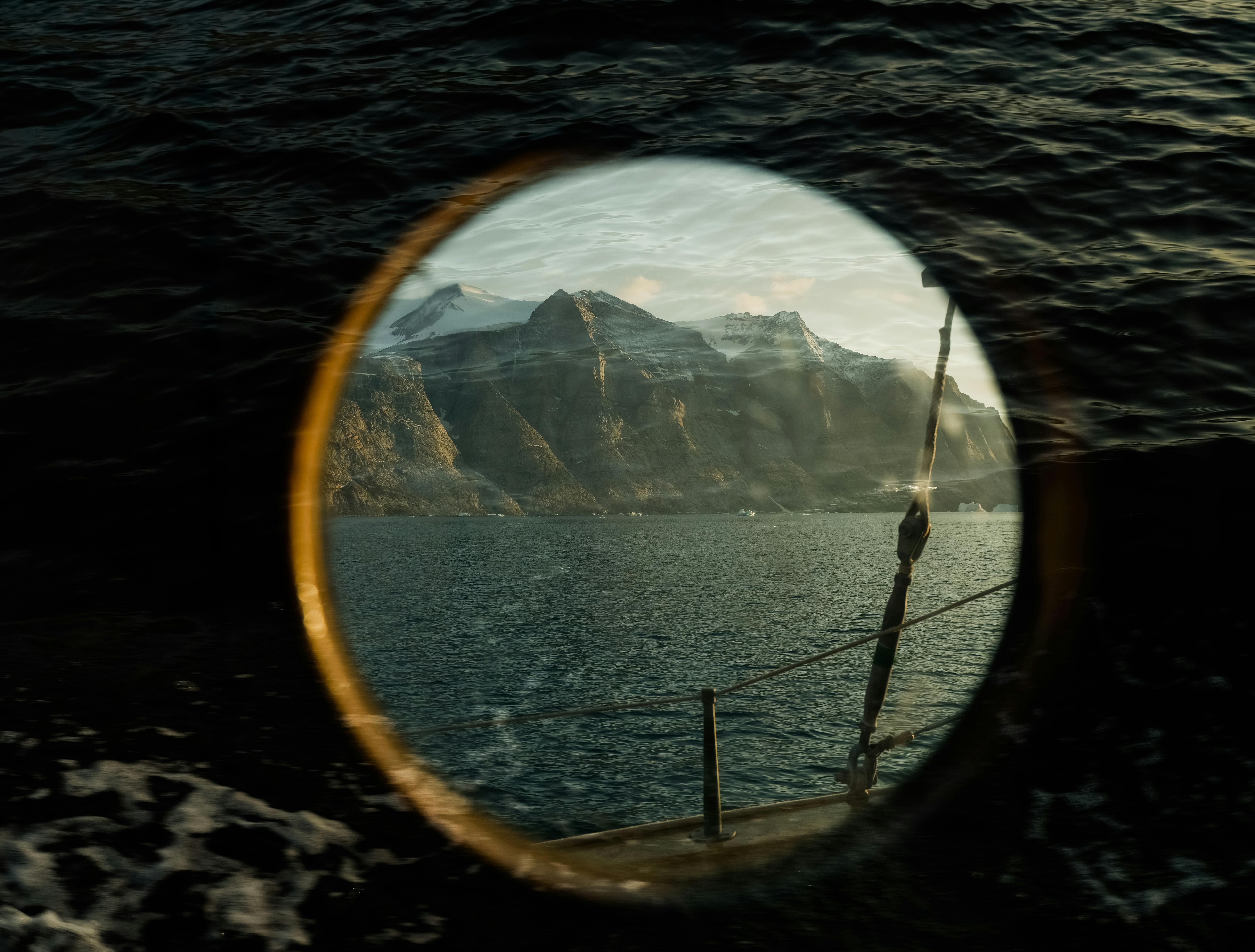 A stunning seascape view through a ship's porthole revealing rugged mountains and deep blue sea.