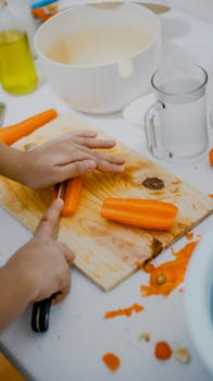 A person chops fresh carrots on a wooden board with a knife, preparing vegetables in the kitchen.