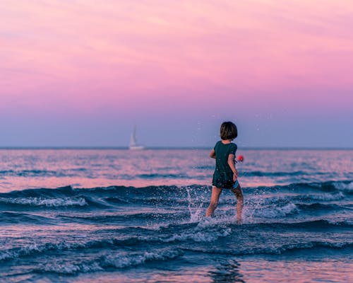 Back view of anonymous kid running along seashore and splashing water while having fun in summer at sundown