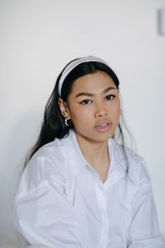 一位戴着耳环、化了妆、身着白衣的年轻少数族裔女性在家中看着镜头