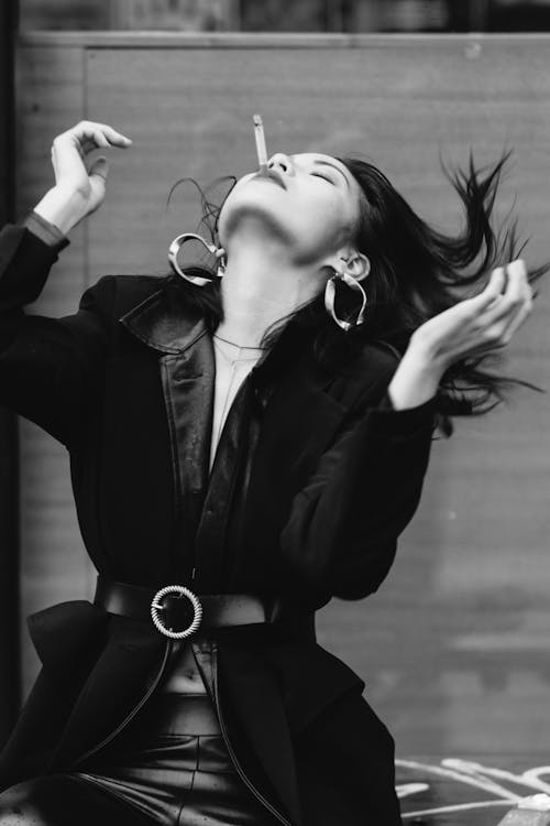 Black and white of trendy ethnic female in earrings with cigarette and closed eyes standing with head thrown back