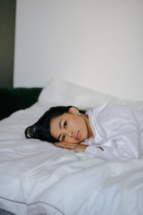 Free Young Asian female in white clothes lying on cozy creased bed sheet and looking at camera at home Stock Photo