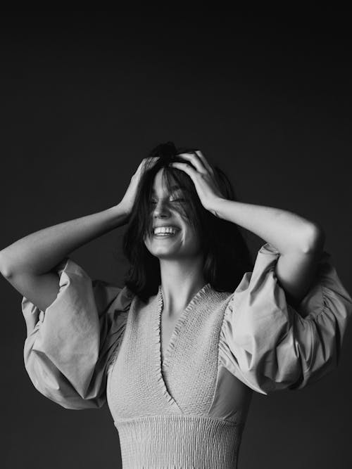 Black and white cheerful young female smiling happily while standing with eyes closed in studio and touching head with both hands