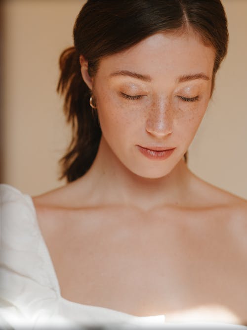 Crop tranquil calm female with freckles wearing white outfit standing with eyes closed against light wall