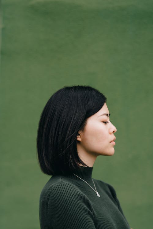 Side view of young dark haired female in casual clothes standing against green background and looking down