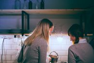 Back view of happy women with dishes standing near sink and having fun together in dark kitchen with light bulb