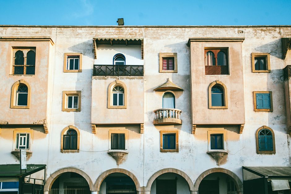 Weathered Mediterranean building facade with sun-bleached, damp-stained walls and aged plaster above arched arcades.