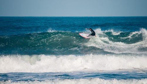 Full body anonymous sporty surfer wearing black wetsuit balancing on surfboard on foamy sea waves against blue sky