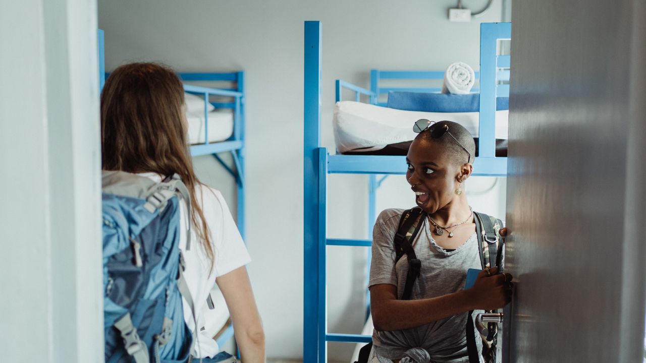 Two backpackers walking into a bright hostel dormitory.