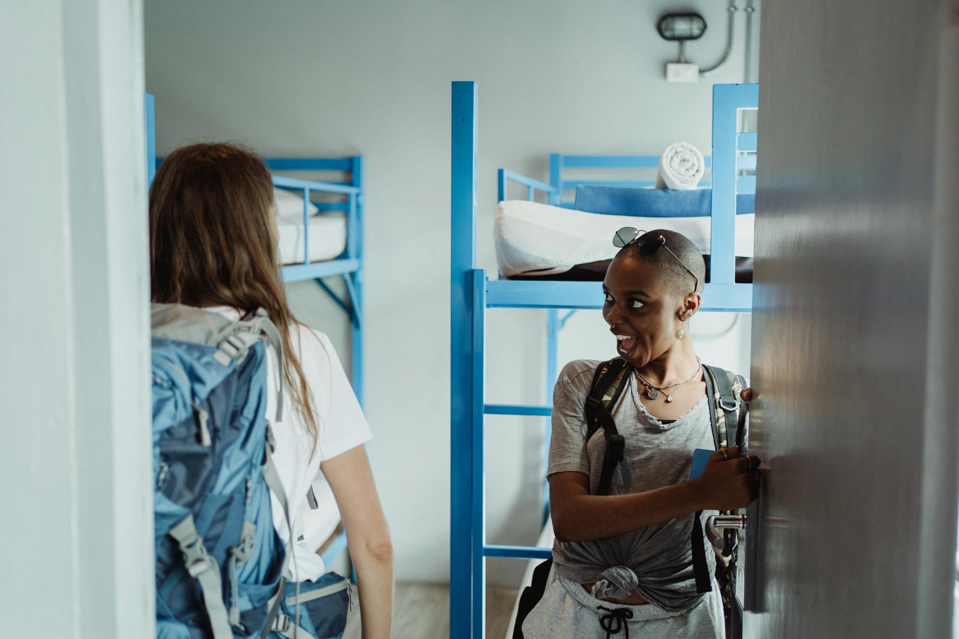 Two women with backpacks walking into a bright hostel dormitory with bunk beds, ready for adventure.