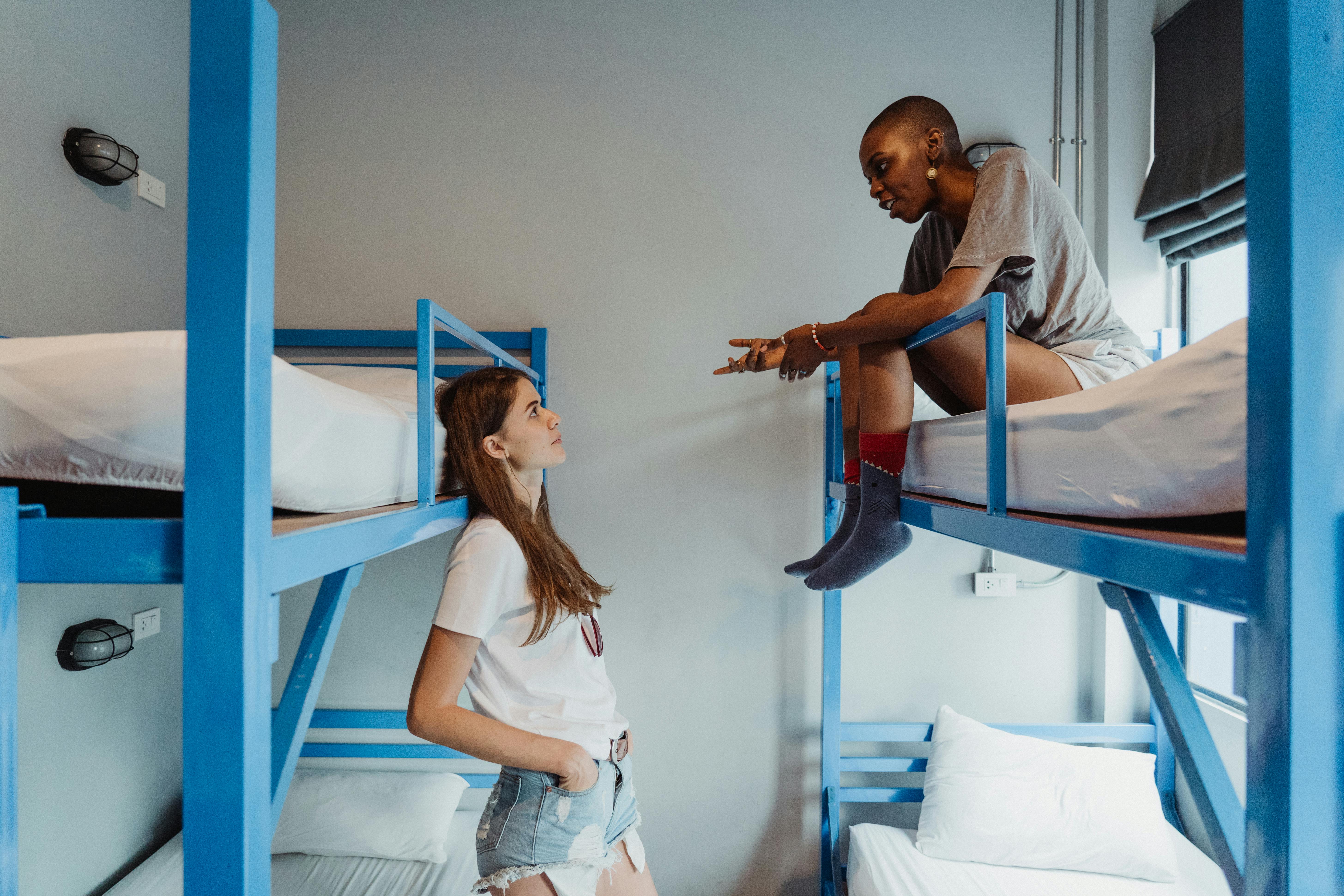 Two women chatting in a hostel dormitory room, with one sitting on a blue bunk bed and the other standing beside it