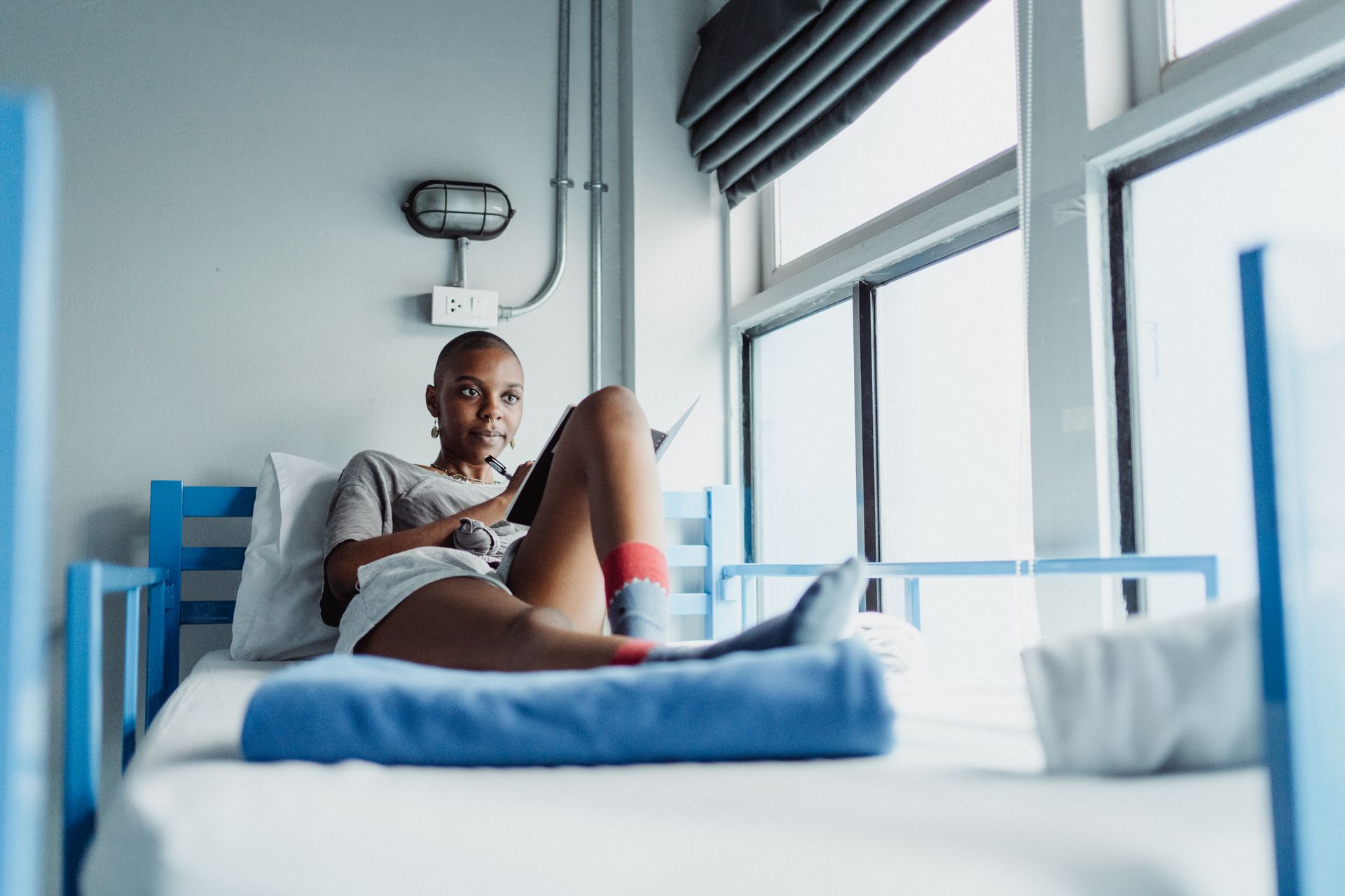 Woman lounging on a bunk bed near a window with a notebook, surrounded by natural light.