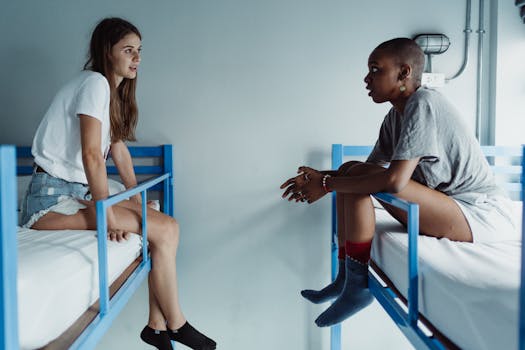 Casual conversation between two women seated on bunk beds in a minimalist room setting.