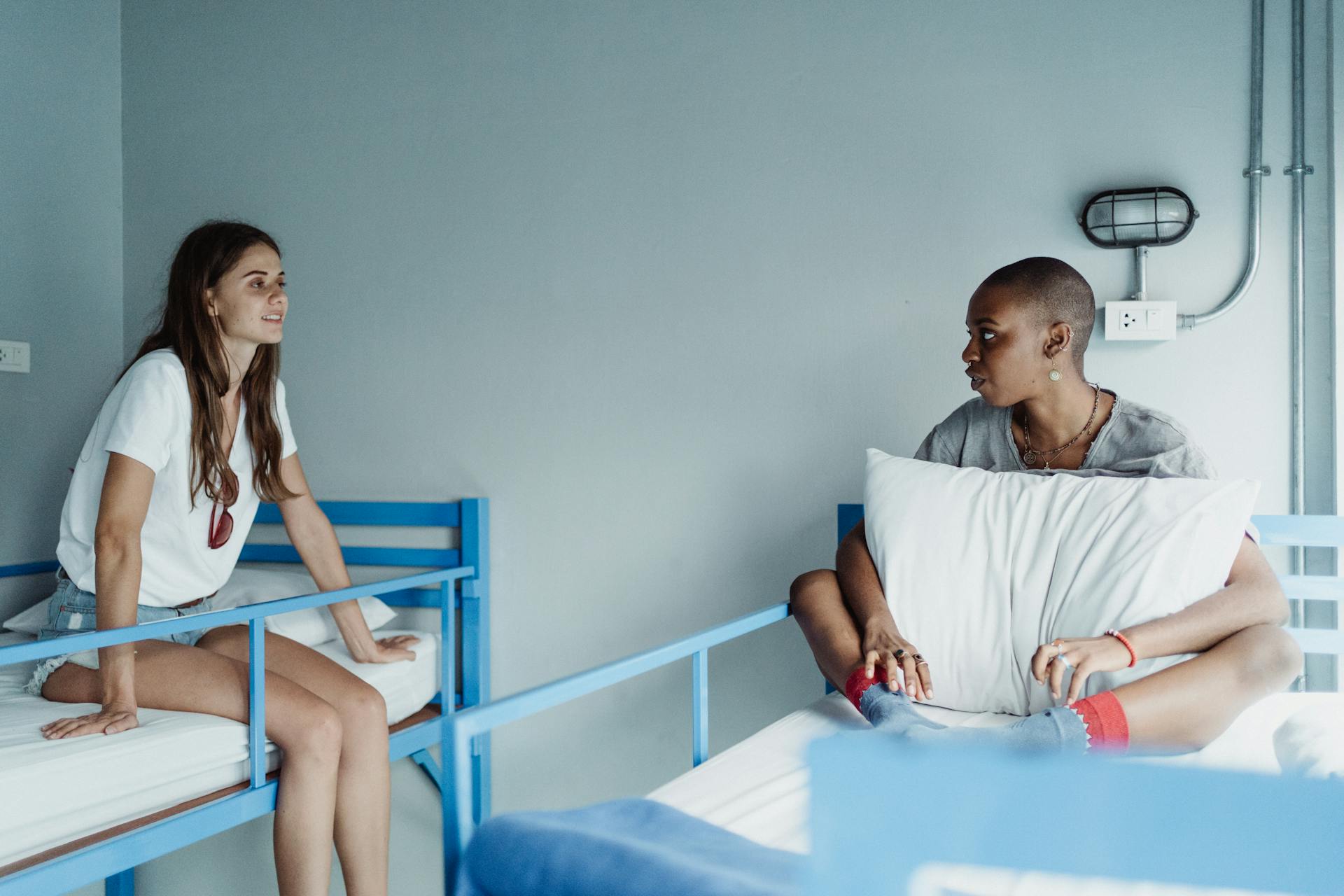Two women sit and converse casually on bunk beds in a hostel room setting.