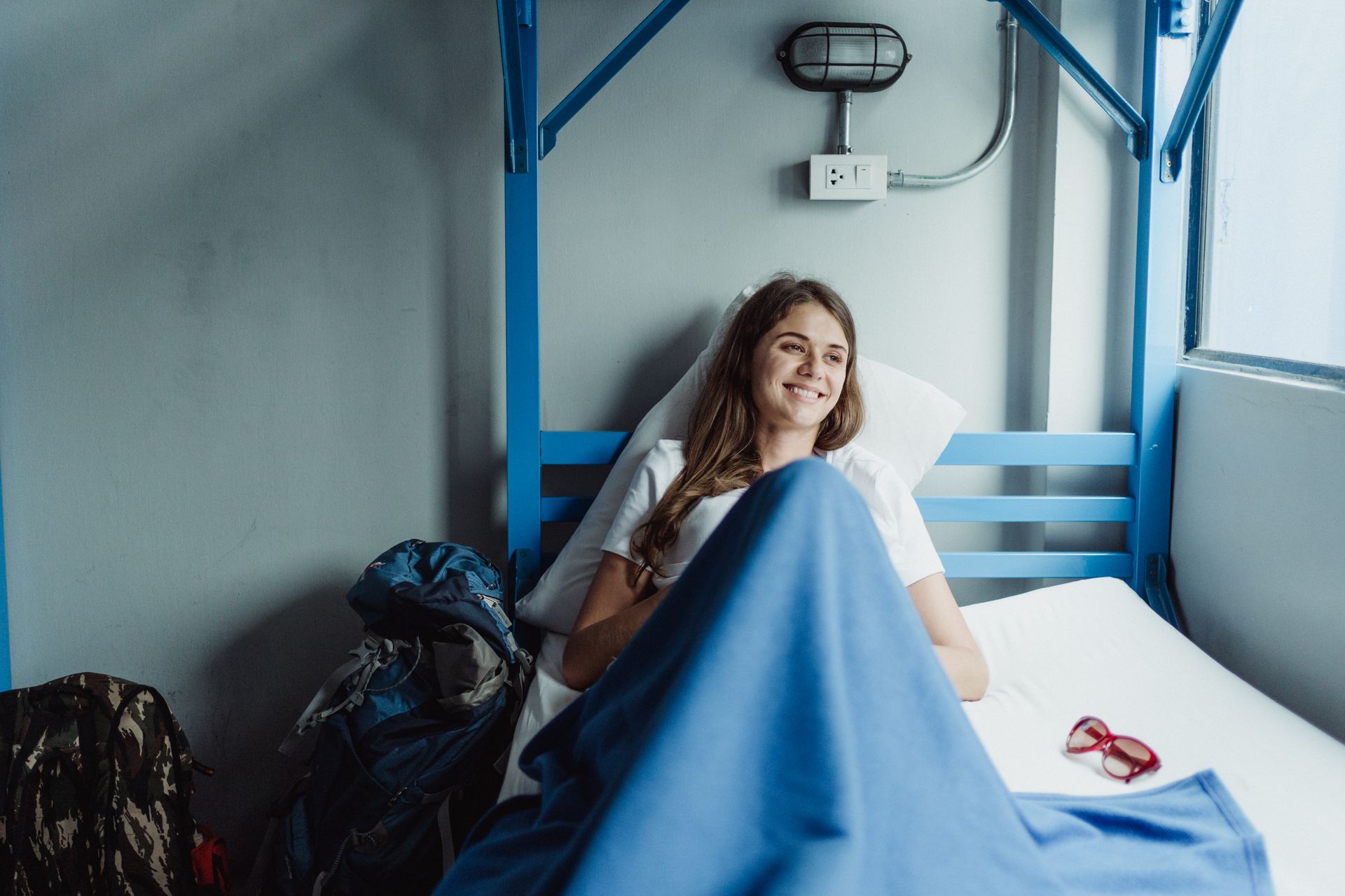 Smiling woman in a bunk bed with backpacks, embracing a relaxing travel experience.