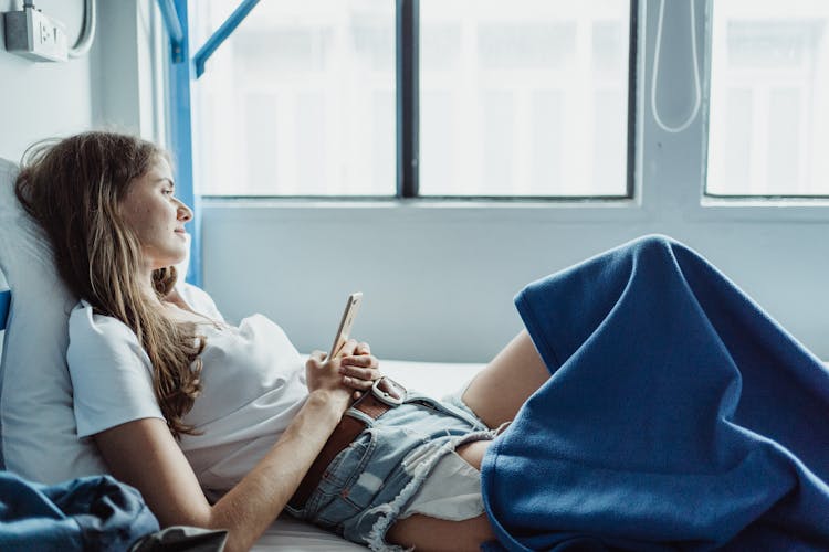 Woman Lying In Bed, Holding A Phone And Looking Out The Window 