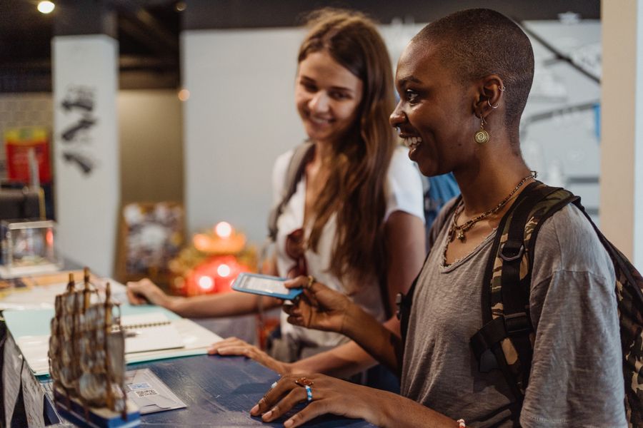 Two smiling travelers with backpacks lean on a reception desk, one holding a blue card, while they check in
