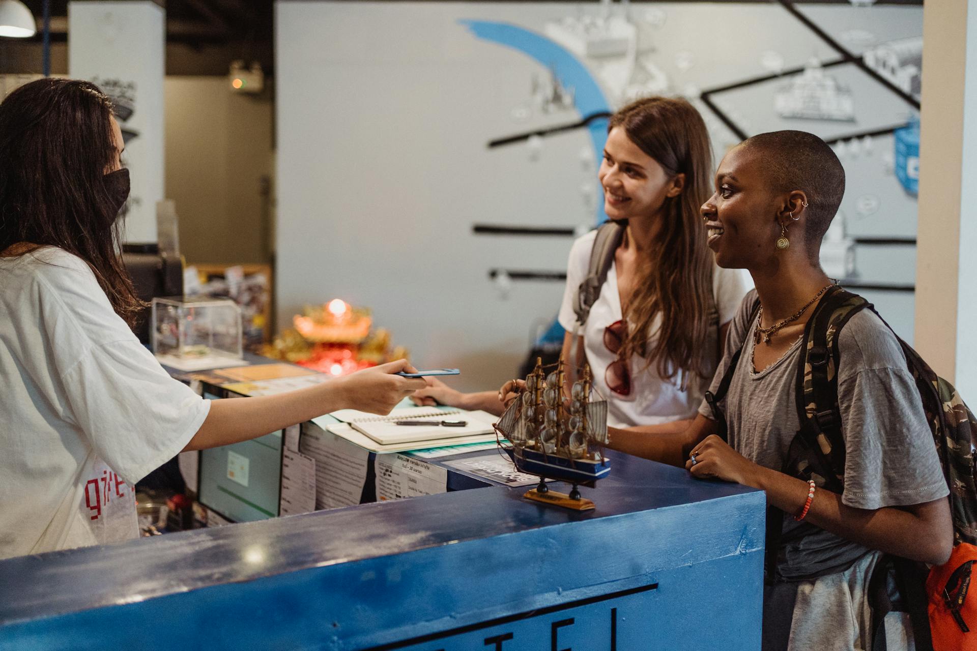 Two women travelers checking in at a hostel reception with a welcoming staff member wearing a face mask.