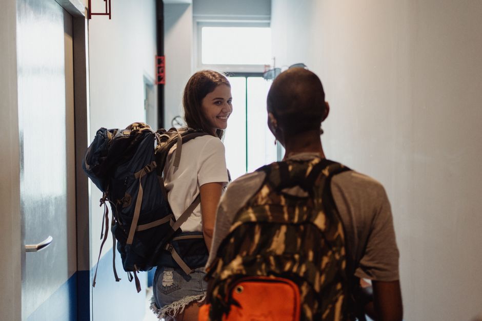 Two young travelers with backpacks leaving a hostel through a narrow corridor.