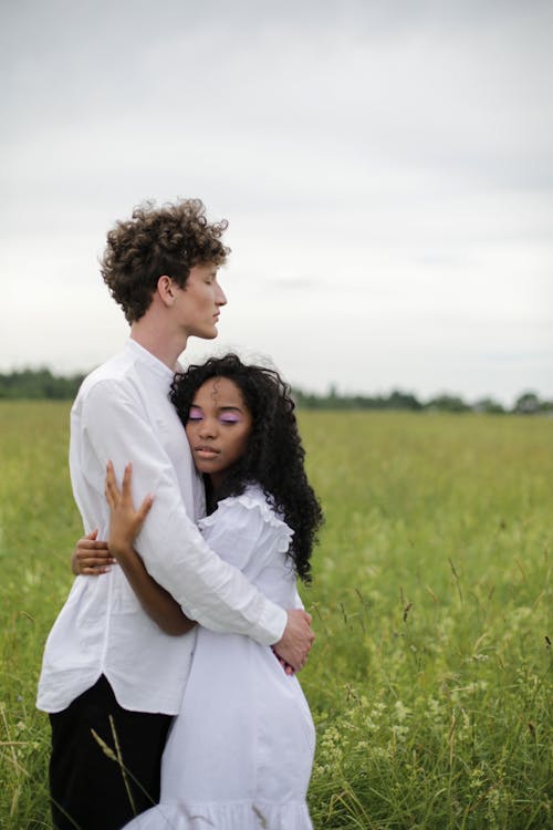 Man in White Dress Shirt Hugging Woman in White Dress on Green Grass Field