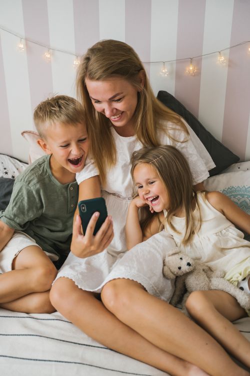 Free A joyful mother and her son and daughter sit on a bed, laughing while looking at a smartphone. Stock Photo