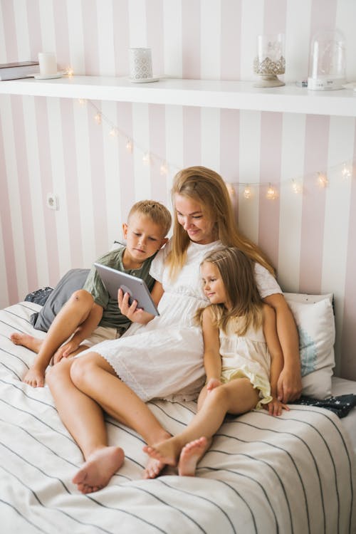 Free Woman and Kids Sitting on Bed While Looking at the Screen of a Tablet Stock Photo