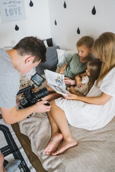Family photoshoot of a man capturing children reading on a cozy bed.