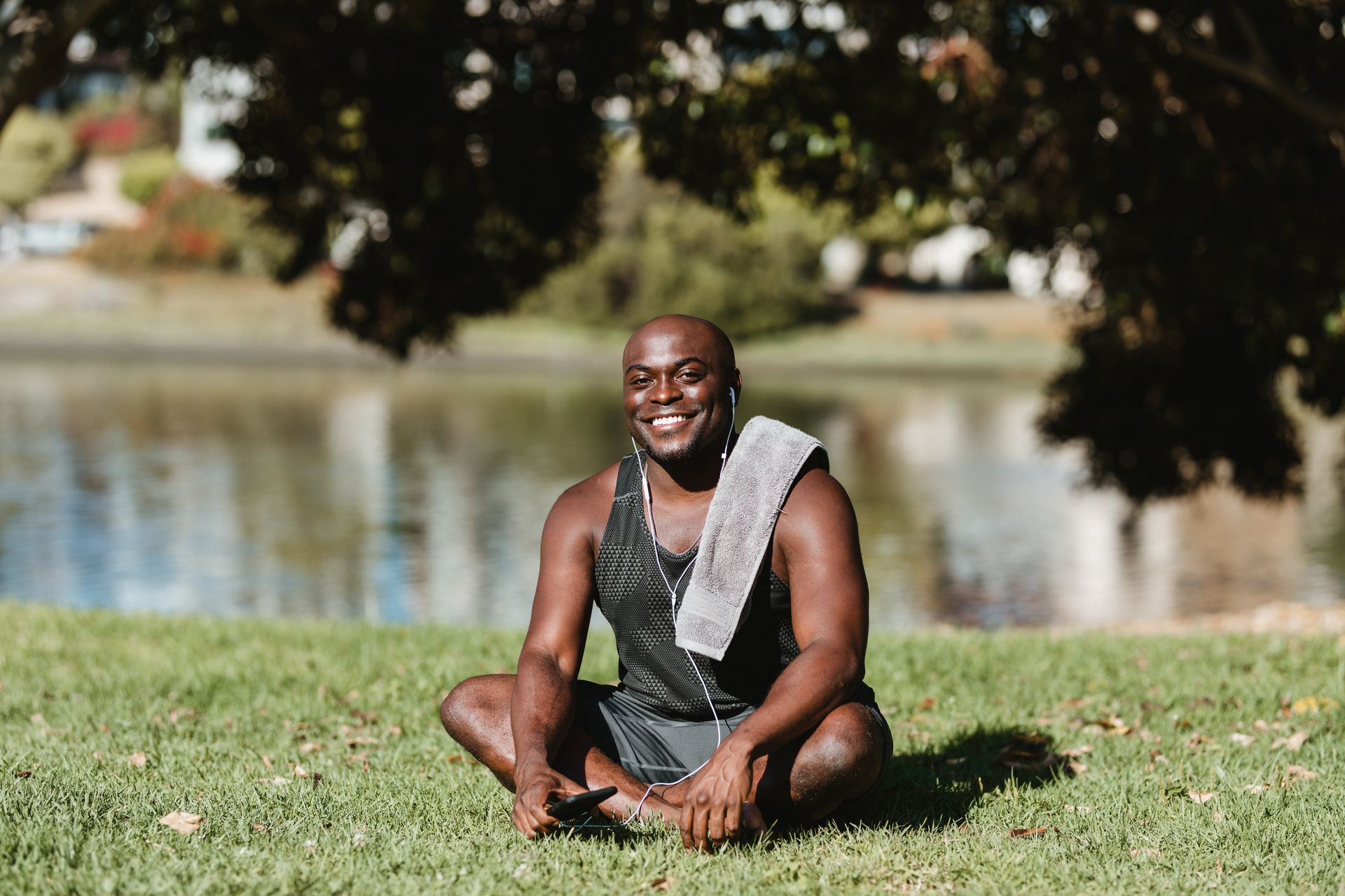 Smiling man with earphones and towel relaxing by a lake in a sunny park setting.