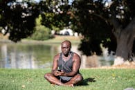 一名成年男子盘腿坐在草地上，戴着耳机用智能手机在户外冥想