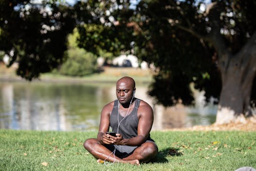 Adult man sitting cross-legged on grass, meditating with smartphone and earphones outdoors.