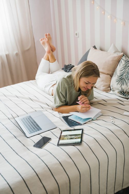 Free Woman writing in a notebook on a bed, surrounded by laptop, smartphone, and tablet, working from home. Stock Photo
