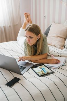 Woman lying on bed working from home using a laptop, tablet, and notebook.