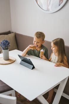 两个孩子在室内一边看平板电脑，一边开心地吃着冰淇淋蛋筒