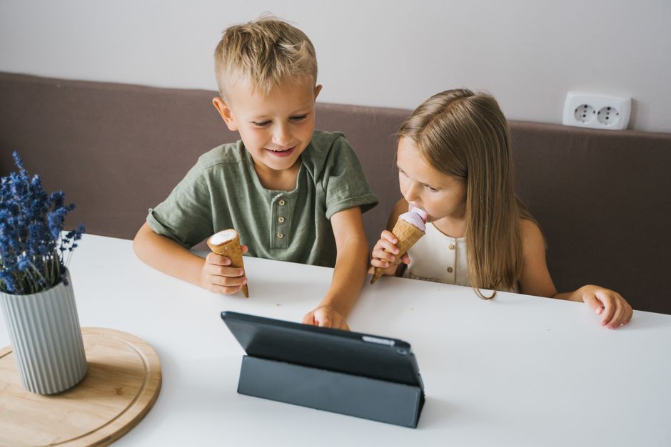 Two children enjoying ice cream cones while watching a tablet indoors.