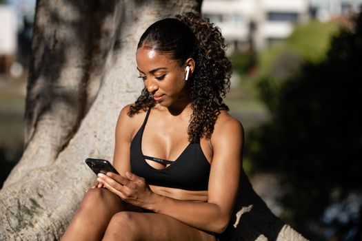 Woman in activewear using smartphone outdoors, sitting on a tree.