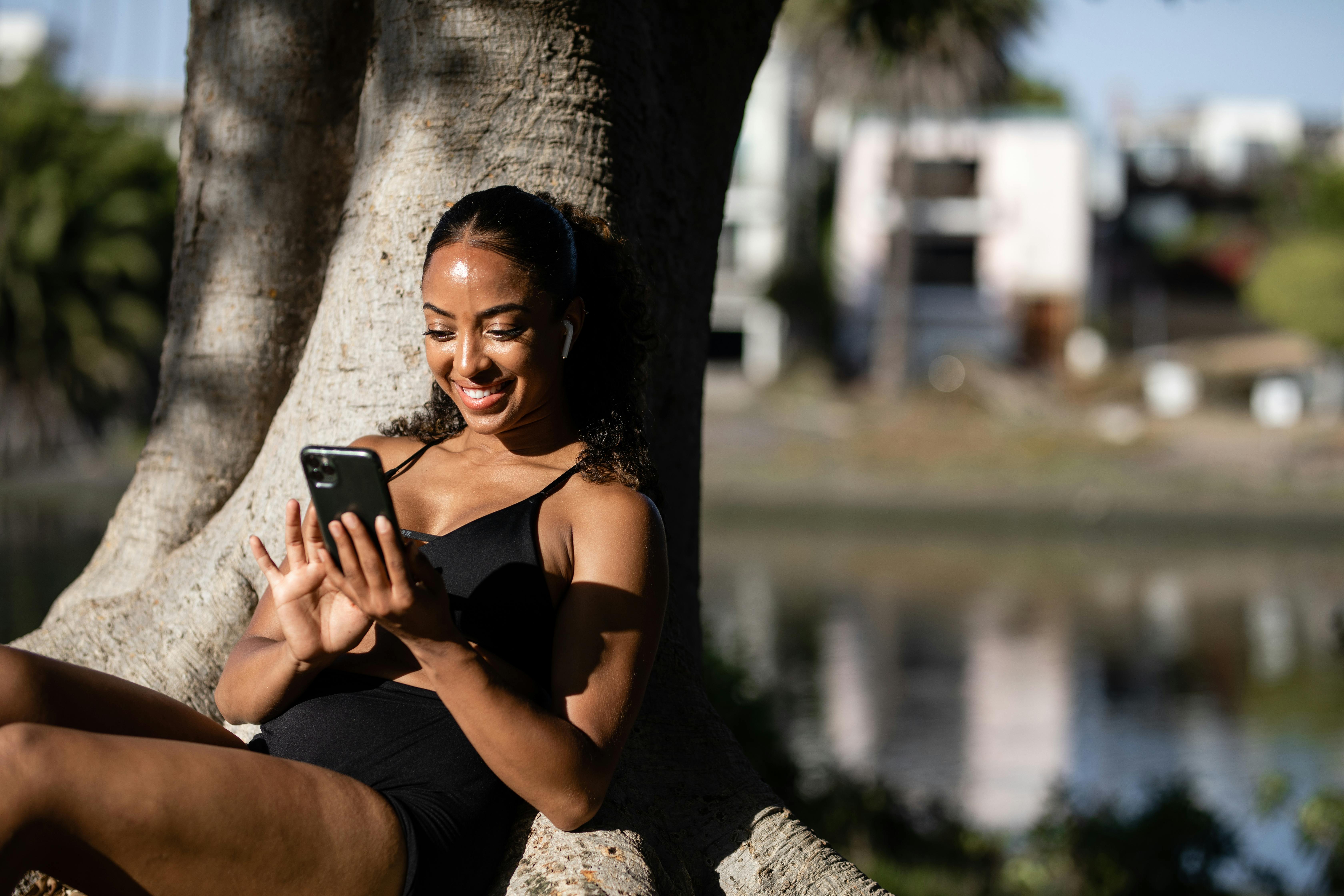 Smiling woman in activewear texting on her phone while leaning against a tree.
