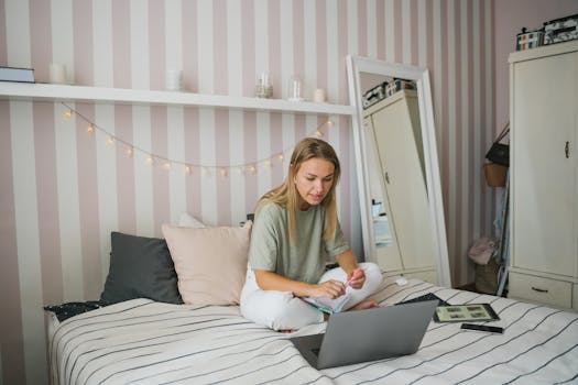 A young caucasian woman working on a laptop while sitting on her bed, with a cozy and relaxed atmosphere in the bedroom.