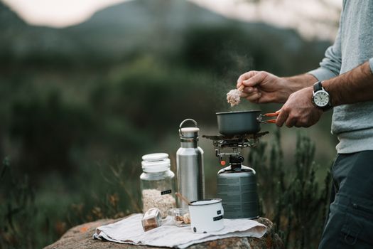Person cooking oatmeal on portable stove in outdoor campsite setting, emphasizing adventure and nature.